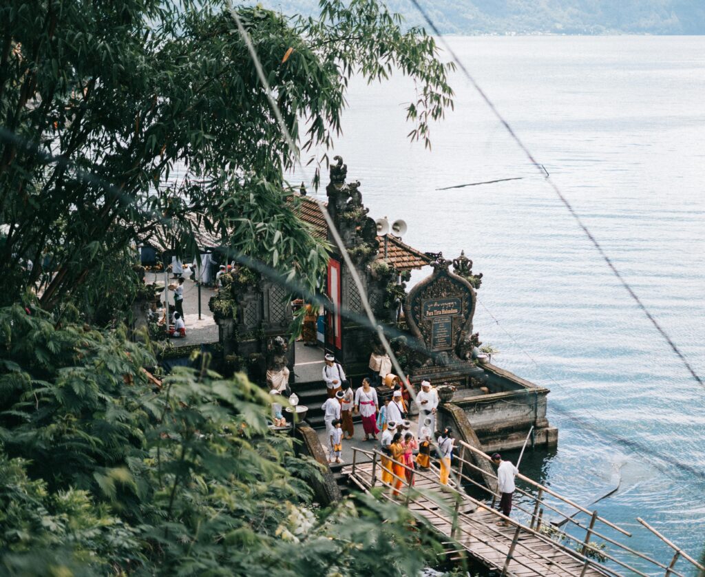 Balinese locals dressed in traditional attire gathering at a lakeside temple in Bali, Indonesia, surrounded by lush greenery and calm water.