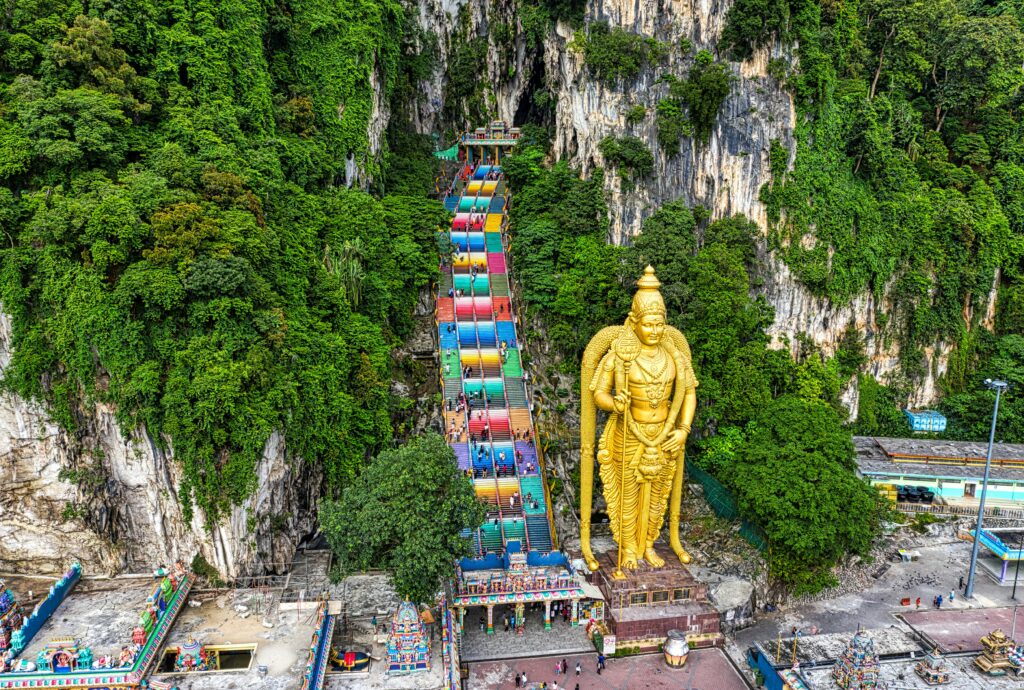 Aerial view of the Batu Caves in Malaysia, featuring the iconic golden Lord Murugan statue and colorful staircase leading into the limestone hills