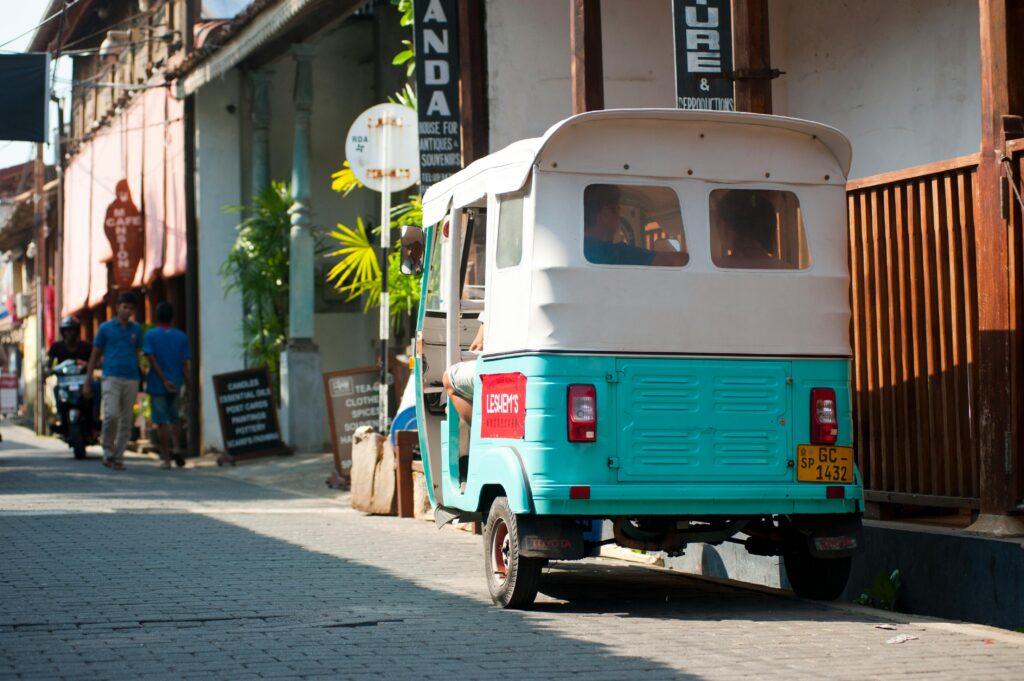 A turquoise and white tuk-tuk parked on a cobblestone street in Galle, Sri Lanka, with locals walking past colorful boutique shops and tropical plants