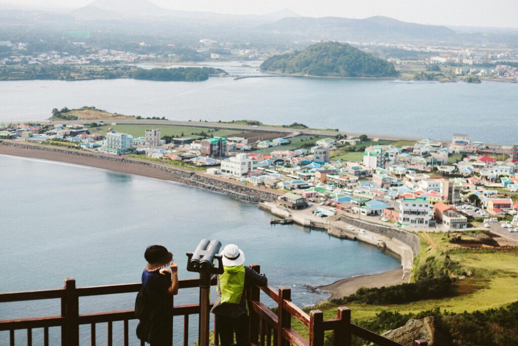 Scenic coastal view of Jeju Island, South Korea, with tourists using binoculars to overlook colorful seaside villages, ocean bay, and lush landscape