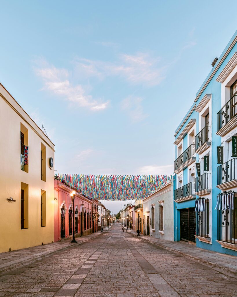 Colorful colonial street in Oaxaca, Mexico with vibrant buildings, hanging decorations, and cobblestone pavement at sunset