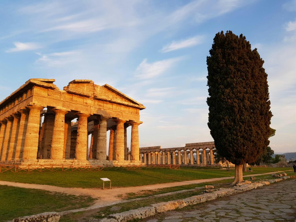 Ancient Greek temple ruins at Paestum, Italy, illuminated by golden sunset light, with a tall cypress tree and stone path in the foreground