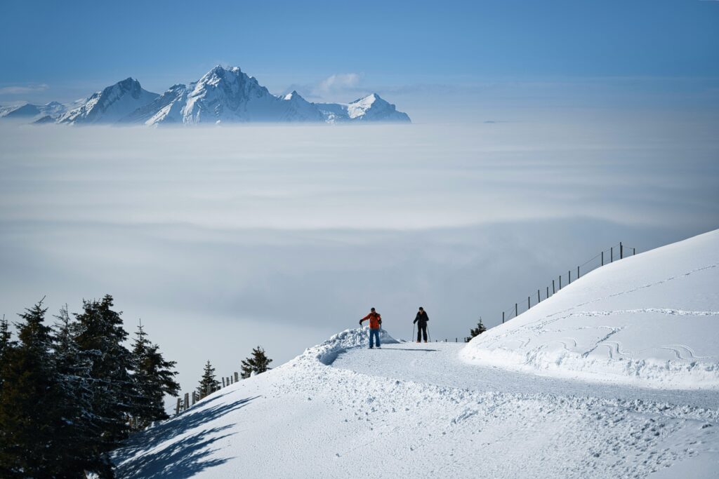 Skiers standing on a snow-covered slope overlooking a sea of clouds with dramatic alpine peaks in the background under a clear blue sky