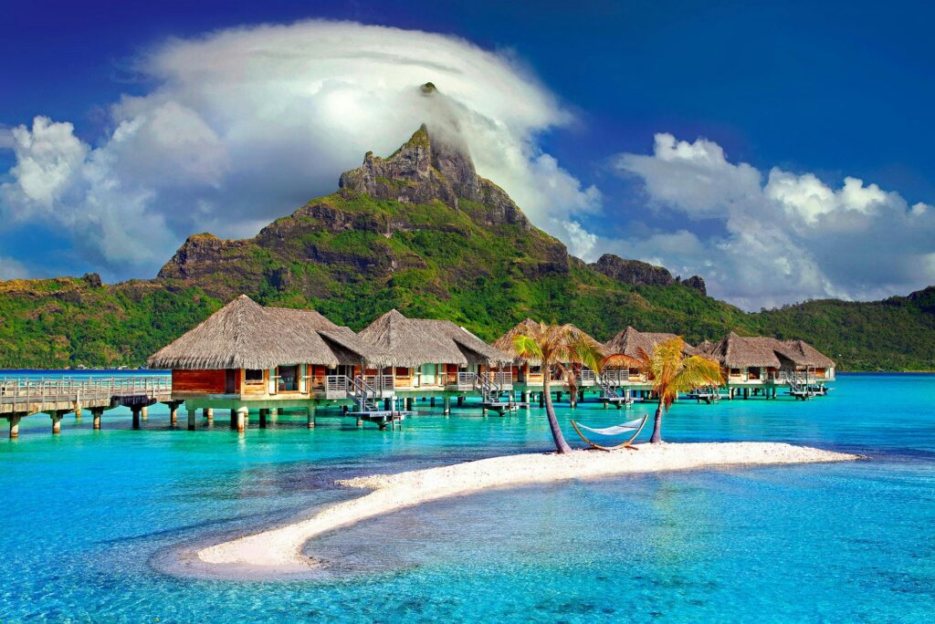 Scenic view of overwater bungalows in Tahiti with turquoise lagoon, white sand islet, and Mount Otemanu in the background under dramatic clouds