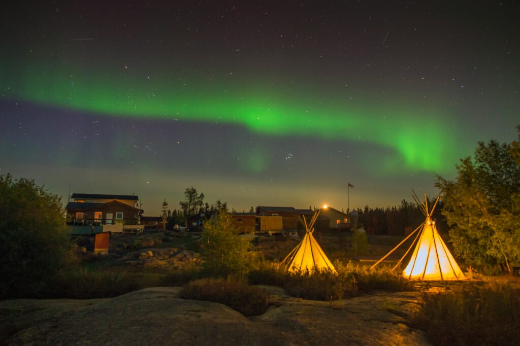Traditional Indigenous teepees glowing under the Northern Lights in Canada, with a starry night sky and forested cabins in the background
