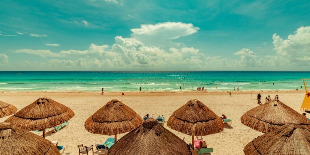 Thatched beach umbrellas and lounge chairs on a sandy beach in Cancún, Mexico, with turquoise ocean waves and people swimming in the background