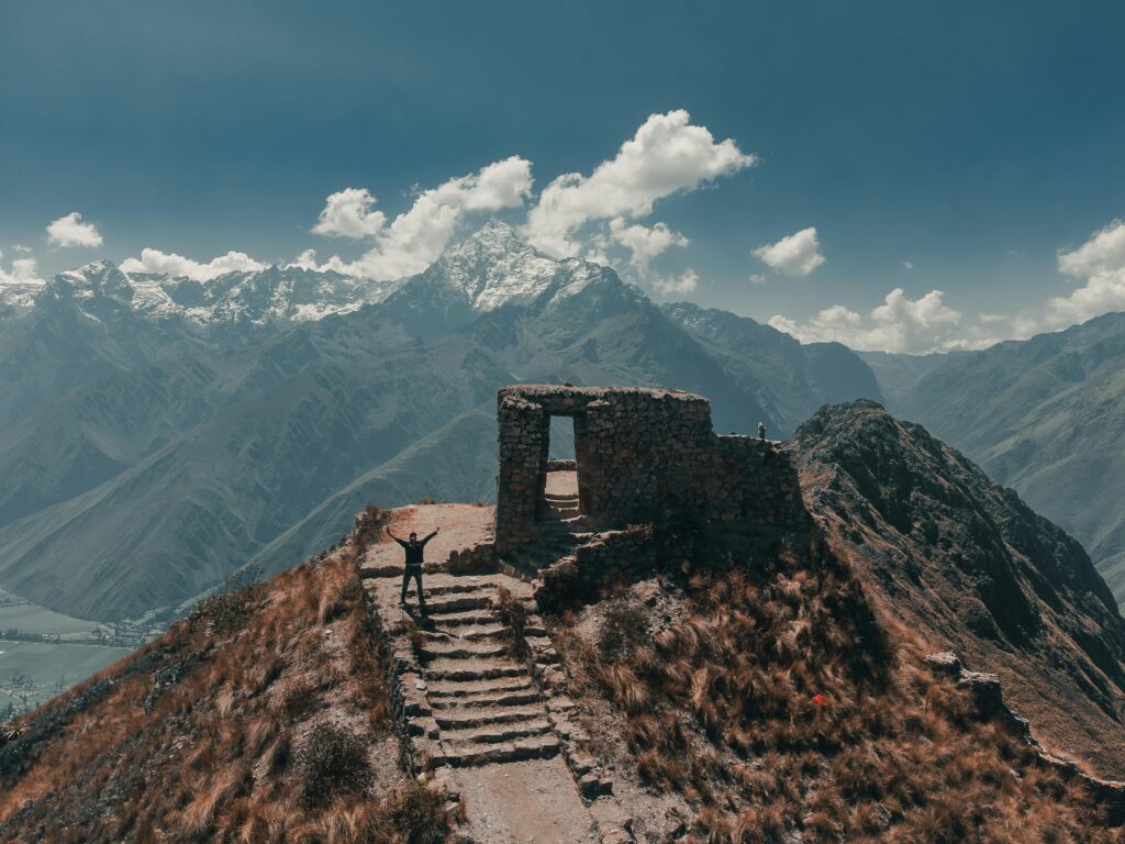 Traveler standing on ancient stone steps beside Inca ruins overlooking the Andes mountains with snow-capped peaks in the Sacred Valley of Peru